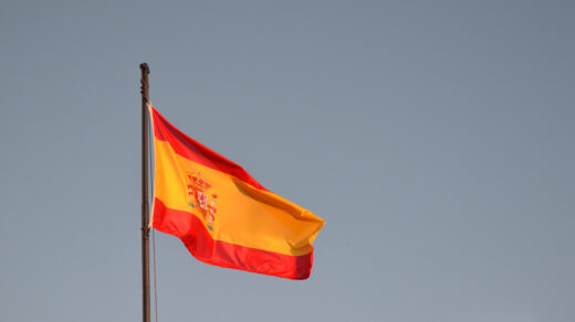 Na malowniczym tle niebieskiego nieba widoczna jest hiszpańska flaga, dumnie powiewająca na wietrze. Jego intensywny czerwony kolor kontrastuje z błękitem nieba, symbolizując dumę, niezależność i bogatą historię Hiszpanii.