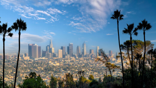 Na tym zdjęciu widoczny jest spektakularny widok na Los Angeles za dnia, z charakterystycznymi wieżowcami w tle i jasnym, bezchmurnym niebem. Widać też rozległe ulice, na których krążą samochody, a w oddali widoczne są malownicze góry.