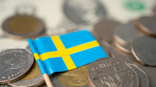 Obrazek przedstawia mianiturową flagę Szwecji, spoczywającą na stosie szwedzkich monet. Symbolika flagi w połączeniu z monetami odzwierciedla zarówno dumę narodową, jak i stabilność gospodarczą kraju, podkreślając znaczenie waluty szwedzkiej w kontekście szwedzkiej tożsamości i ekonomii.