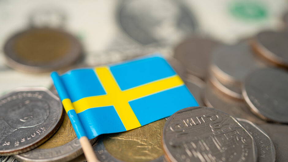 Obrazek przedstawia mianiturową flagę Szwecji, spoczywającą na stosie szwedzkich monet. Symbolika flagi w połączeniu z monetami odzwierciedla zarówno dumę narodową, jak i stabilność gospodarczą kraju, podkreślając znaczenie waluty szwedzkiej w kontekście szwedzkiej tożsamości i ekonomii.