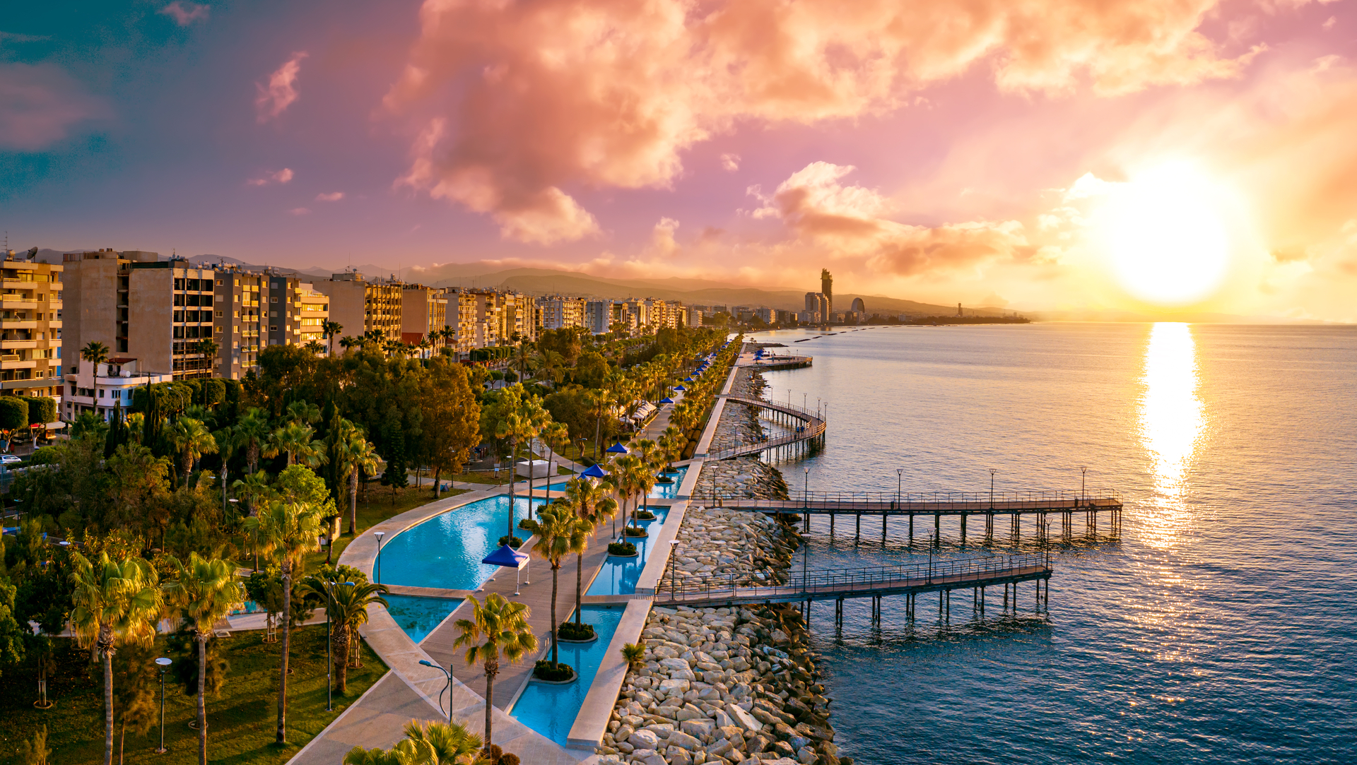 Na tym obrazku przedstawiono piękny krajobraz Cypru, z piaszczystą plażą i luksusowymi hotelami, które wyrastają zielonymi ogrodami. W oddali widoczne są także góry, które dodają tej scenerii jeszcze więcej uroku. Plaża jest idealnym miejscem do relaksu i wypoczynku, a hotele oferują wysoki standard usług, aby zapewnić swoim gościom wygodę i komfort podczas ich pobytu na wyspie.