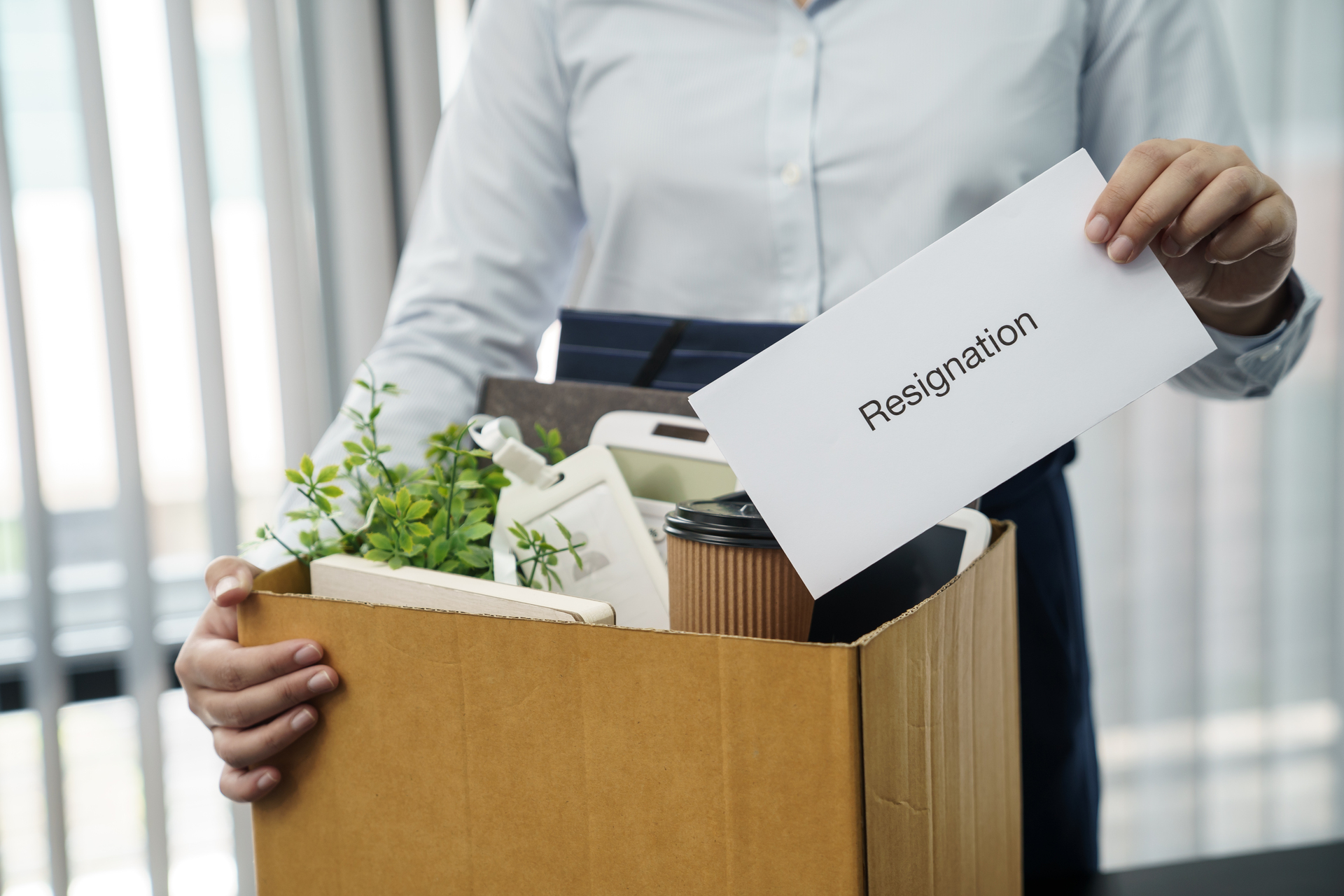 Kobieta wyprowadzająca się z biura z pudłami i białą kratką w ręce z napisem "resignation"