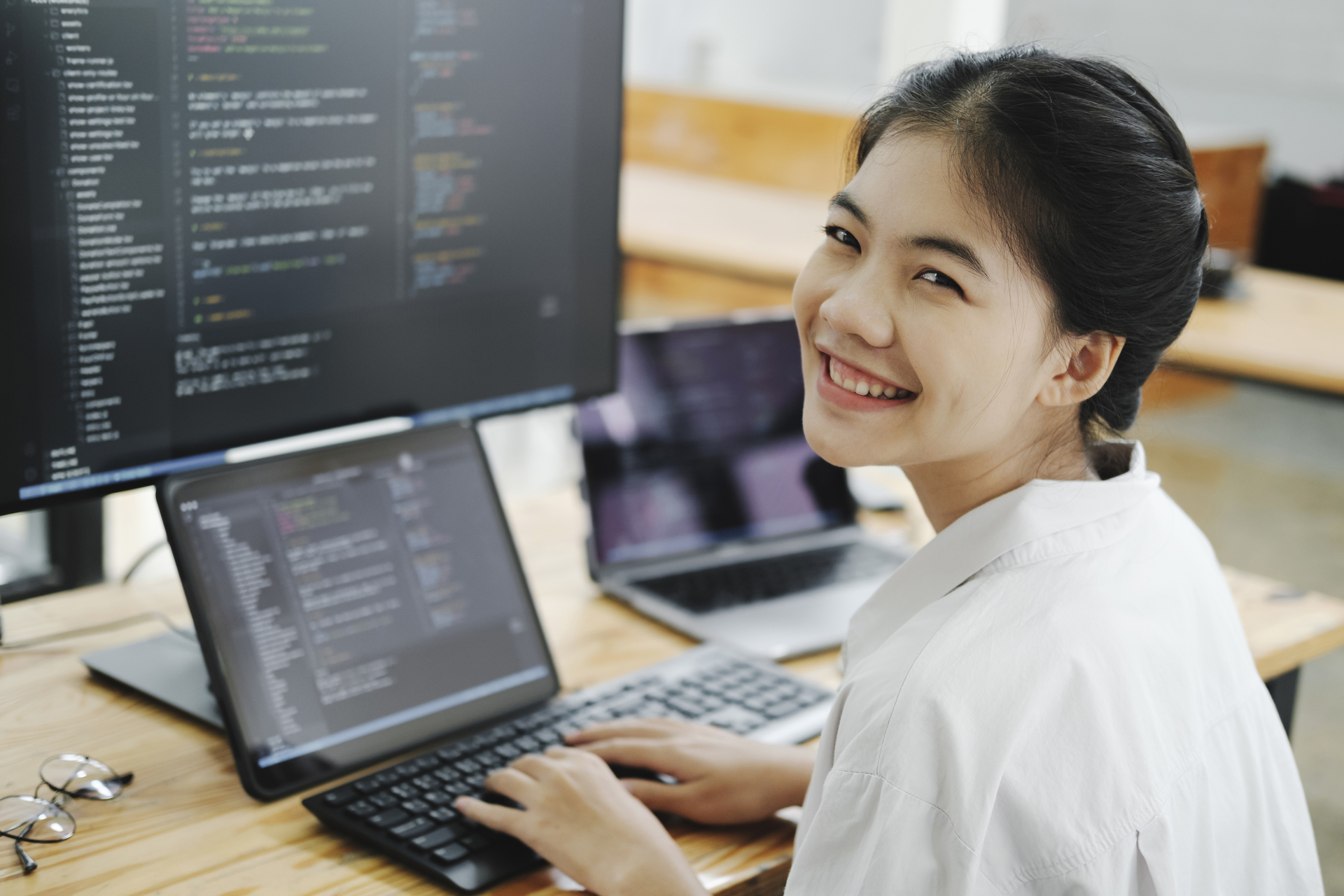 Programistka, siedząca przed komputerem, otoczona monitorami i uśmiechająca się do aparatu